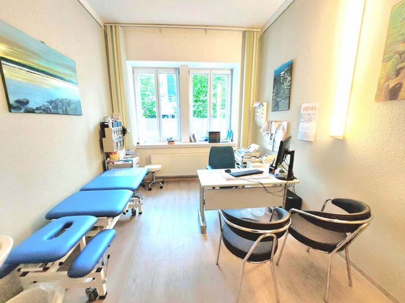 Heller Behandlungsraum mit blauer Behandlungsliege, Schreibtisch am Fenster und schwarzen Besucherstühlen