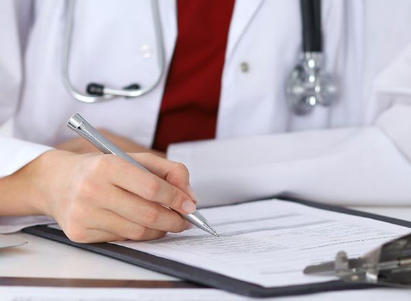 Arzt in weißem Kittel mit Stethoskop schreibt mit Kugelschreiber auf Dokumente in einer Klemmbrettmappe am Schreibtisch