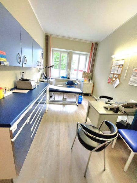 Moderner Behandlungsraum einer Arztpraxis mit blauen Schränken, Behandlungsliege und hellem Tageslicht durch das Fenster