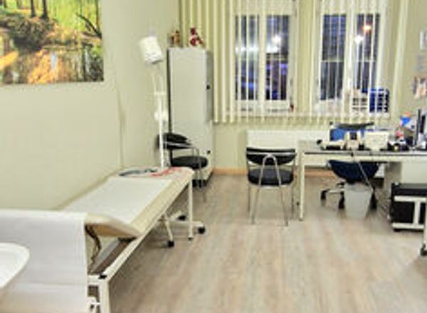 Heller Behandlungsraum mit weißer Behandlungsliege, Schreibtisch am Fenster und Waldbild an der Wand